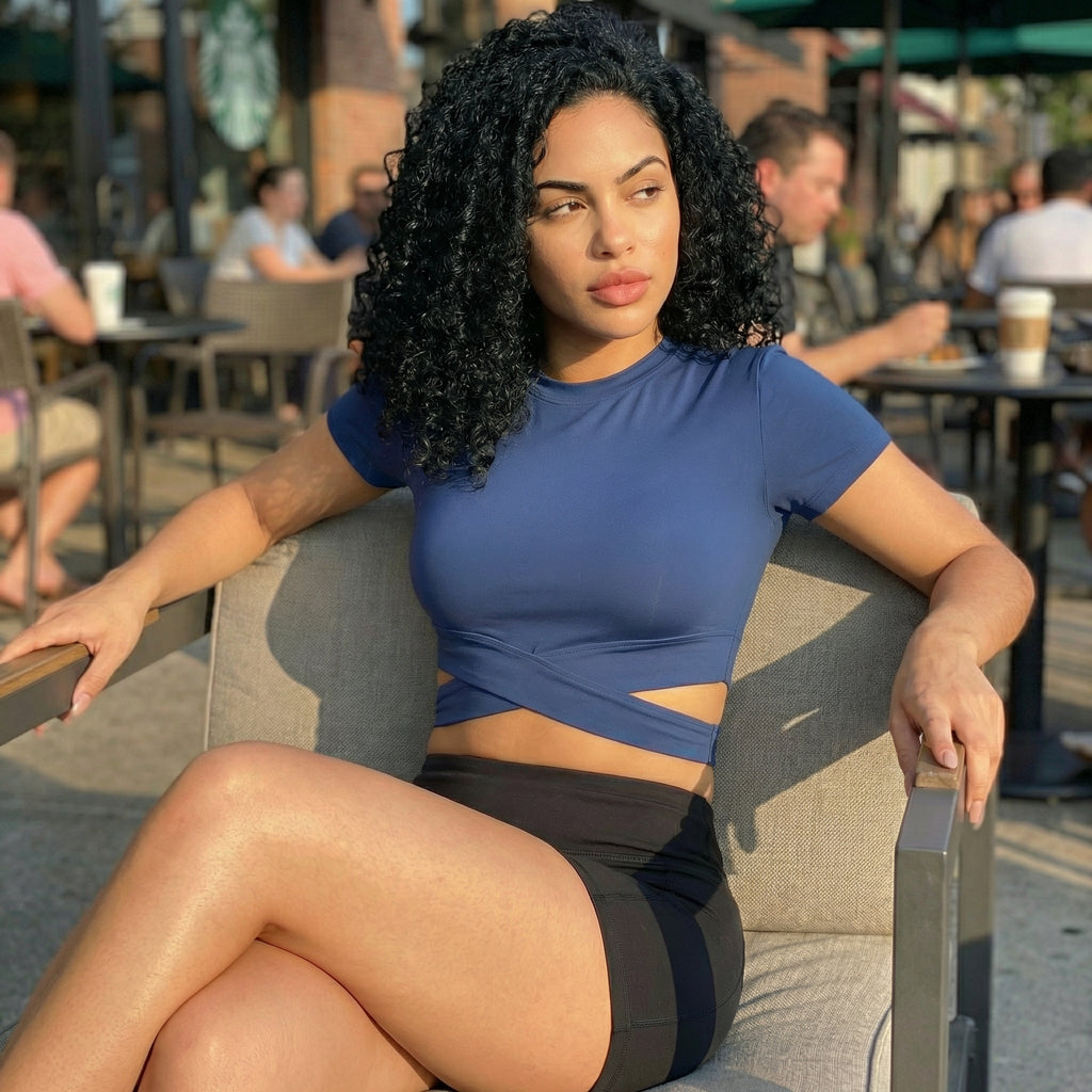 Woman in a blue top and black shorts sitting on a chair outdoors with people and tables in the background.