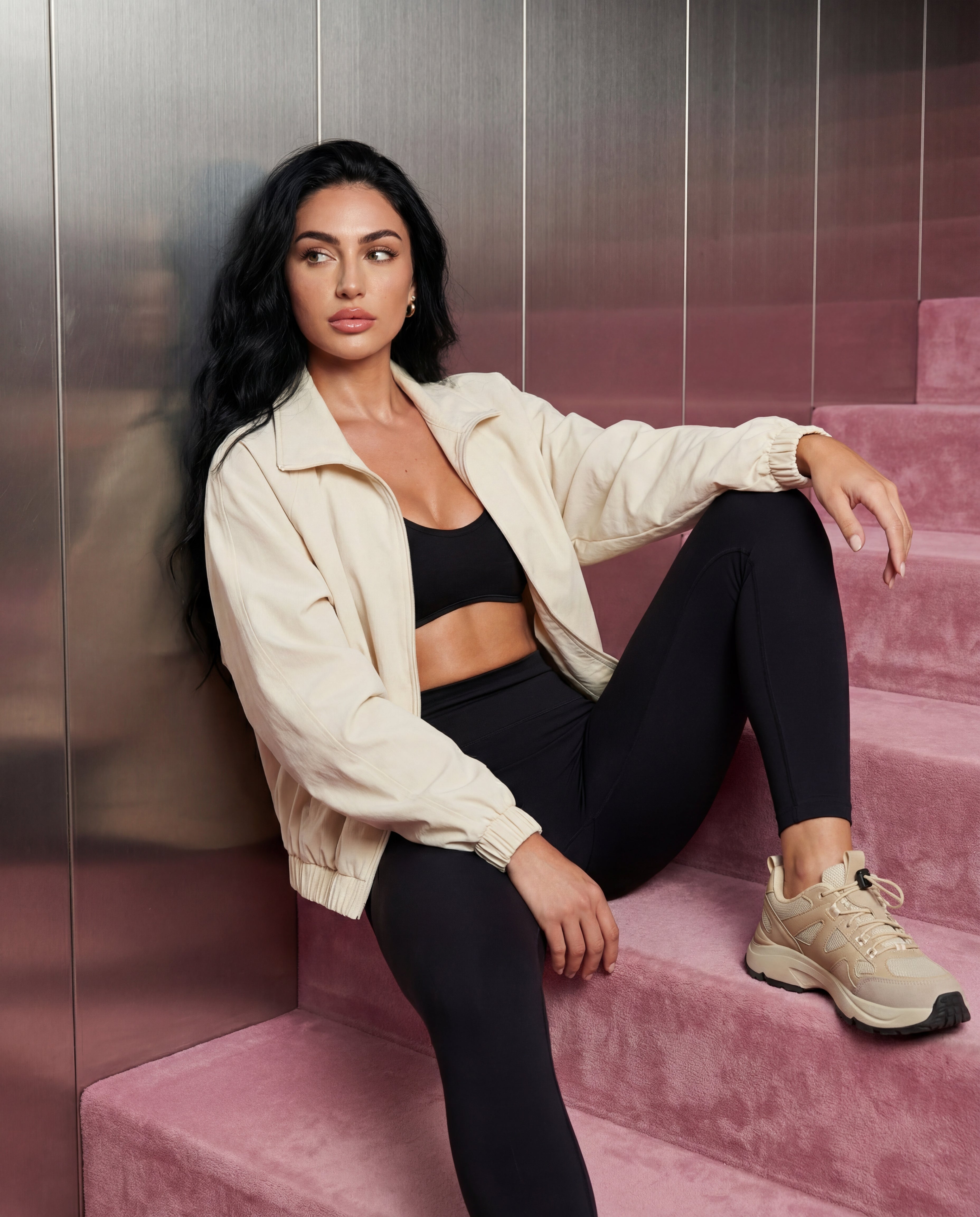 Woman sitting on pink steps wearing a beige jacket, black sports bra, and leggings.