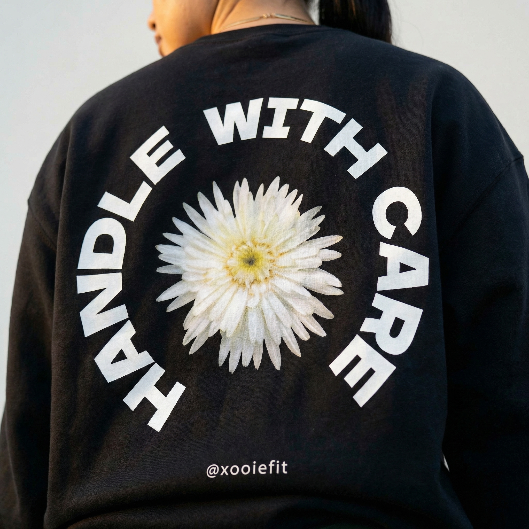 Black crewneck sweatshirt back close-up: white daisy with circular "WITH CARE" text